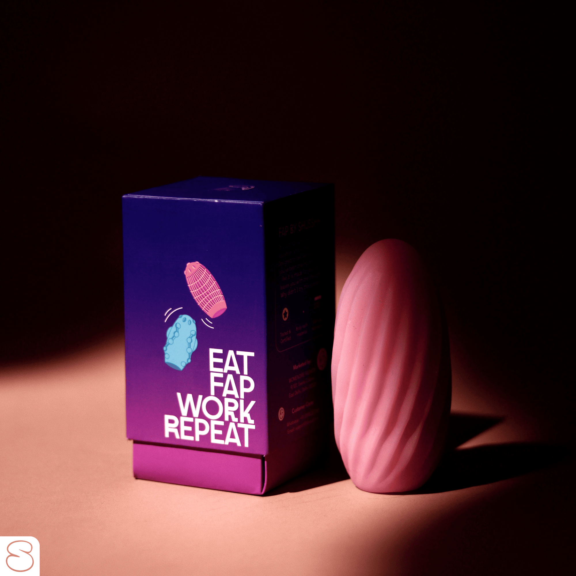 Fap stroker in pink color placed beside its purple packaging box with 'Eat Fap Work Repeat' text, shot in soft studio lighting